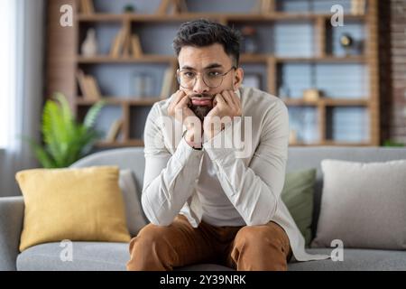 Jeune homme avec des lunettes assis sur le canapé, profondément dans la pensée, réfléchissant sur les choix de vie. Habillé dans des vêtements décontractés, véhiculant introspection et contemplation dans un cadre de détente à la maison. Banque D'Images