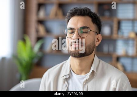 Photo en gros plan d'un jeune homme indien en bavoir et chemise à la maison, assis les yeux fermés, rêvant et se reposant. Banque D'Images