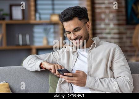 Homme souriant à l'aide d'un smartphone sur le canapé, profiter du temps libre à la maison. Capture la joie, la connectivité et la détente avec la technologie moderne. Représentation parfaite du style de vie numérique Banque D'Images