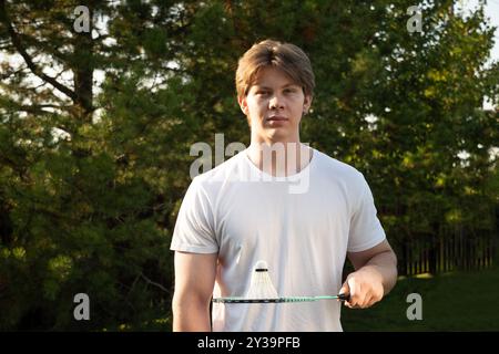 Jeune homme en t-shirt blanc tenant un volant de badminton blanc et une raquette de badminton. Jeu d'été en plein air. Banque D'Images