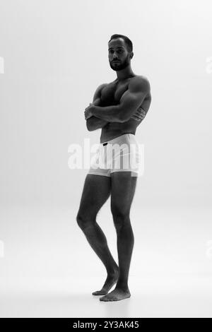Homme musclé se tient confiant dans un boxer blanc, mettant en valeur son physique tonique sur fond blanc. Filtre monochrome. Banque D'Images