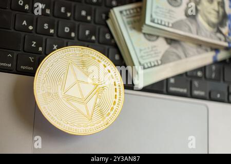 KIEV, UKRAINE - 17 AOÛT 2024 Ethereum ETH coin avec une grande quantité de billets de cent dollars américains sur la surface de l'ordinateur portable en gros plan Banque D'Images