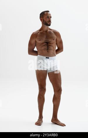 L'homme se tient debout avec ses mains serrées derrière son dos, sa position ferme et confiante respire le contrôle et le calme sur fond blanc. Banque D'Images