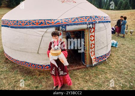 Femme kirghize vêtue de vêtements traditionnels tenant du pain devant une yourte décorée avec des gens en arrière-plan. Banque D'Images