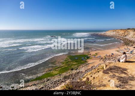 Femme assise au sommet d'une falaise donnant sur l'océan Atlantique et Matadouro Beach à Ericeira à marée basse par une journée venteuse. Portugal Banque D'Images