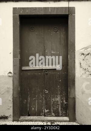 Une image monochrome sépia d'une vieille porte en bois vieillie clouée avec une barre en bois et de la peinture écaillée sur un mur extérieur Banque D'Images