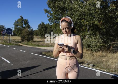 Jeune femme femme écoutant de la musique sur une route asphaltée par une journée ensoleillée, Runner femme écoutant de la musique avec des écouteurs sur une route asphaltée. Concept de RU Banque D'Images