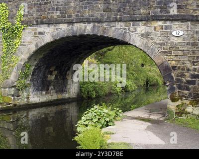 Un vieux pont de pierre traversant le canal de rochdale à mytholmroyd West yorkshire avec un sentier au bord de l'eau entouré d'arbres et de buissons Banque D'Images