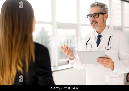 Dans un bureau bien éclairé avec vue sur la fenêtre, un médecin mature aux cheveux gris se tient confiant, les bras croisés, dépeignant professionnalisme et expérience dans le domaine médical. Banque D'Images