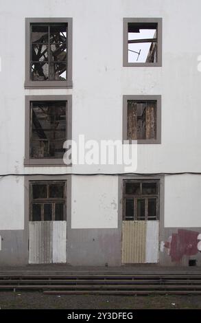 La façade d'un grand bâtiment industriel blanc abandonné avec des portes arborées et des fenêtres vides étayant l'intérieur sans toit accidenté Banque D'Images