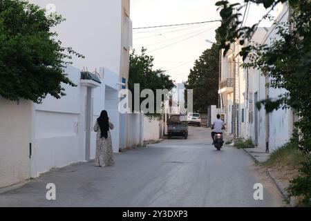 Une femme se tient debout comme un homme qui monte un scooter dans une rue latérale de la ville tunisienne de Hammamet dans le soleil de la fin de l'après-midi Banque D'Images