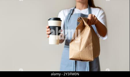 Nourriture et boissons à emporter. Une femme dans un tablier bleu tient une tasse de café de voyage et un sac en papier recyclé avec de la nourriture Banque D'Images