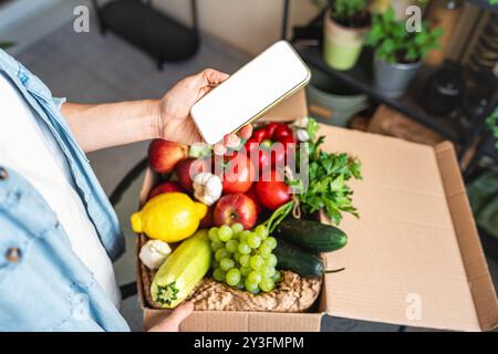 Application mobile Mockup commandant des légumes cultivés à la ferme. Téléphone portable avec écran blanc dans la main féminine au-dessus de la boîte avec des légumes biologiques frais et mûrs. Banque D'Images