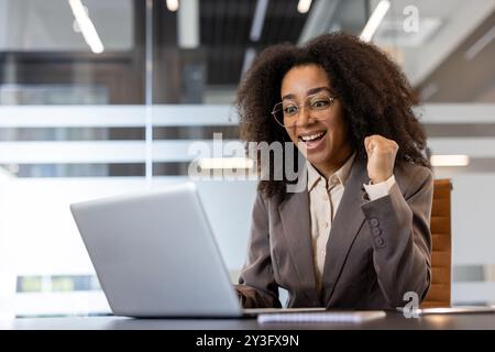 Jeune femme d'affaires afro-américaine aux cheveux bouclés assise en costume dans le bureau à table, regardant l'ordinateur portable, et heureuse du succès montrant le geste de victoire avec la main et disant oui. Banque D'Images