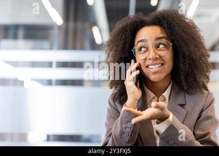 Photo en gros plan d'une jeune femme afro-américaine souriante assise à son bureau, regardant loin et parlant au téléphone portable. Banque D'Images