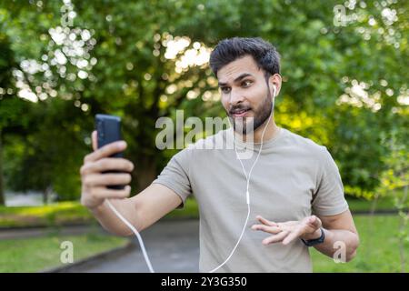 Jeune homme dans une tenue décontractée ayant un appel vidéo énergique dans le cadre du parc. Tenir un smartphone, porter des écouteurs, exprimer des émotions et des gestes engageants. Capturer l'essence de la communication moderne Banque D'Images