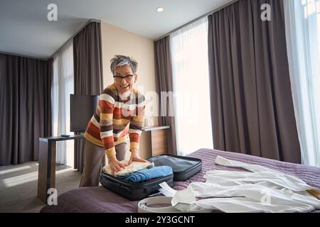 Femme senior positive emballant des vêtements dans la valise dans la chambre d'hôtel Banque D'Images