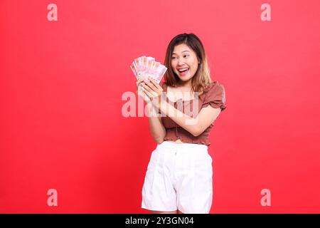 l'expression de la femme d'indonésie est joyeuse, heureuse, franche, les deux mains tenant de l'argent rupiah, portant une blouse brune, fond rouge. pour la transaction, tec Banque D'Images