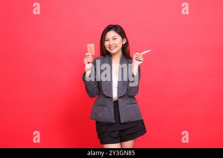 Joyeux bureau indonésie expression de femme tenant une carte de crédit de débit tout en pointant vers la gauche portant une veste et une jupe sur un fond rouge. fo Banque D'Images