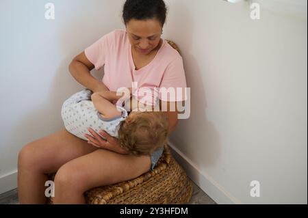 Une mère allaitant tendrement son bébé assis dans une pièce confortable. L'image capture la chaleur de l'affection maternelle et le lien intime entre eux Banque D'Images