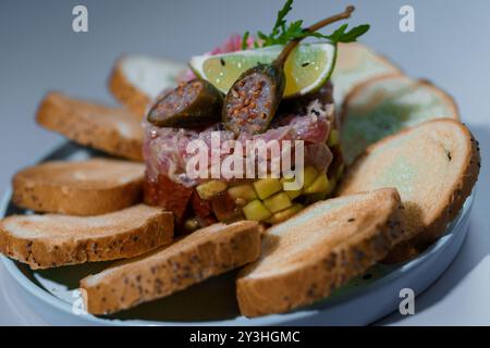 Tartare de thon gastronomique exquis avec avocat et câpres garni de citron vert et d'Arugula, entouré de pain grillé sur une assiette élégante. Banque D'Images