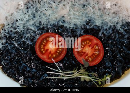 Présentation culinaire exquise avec risotto noir, tranches de tomates juteuses et délicate garniture aux herbes. Banque D'Images