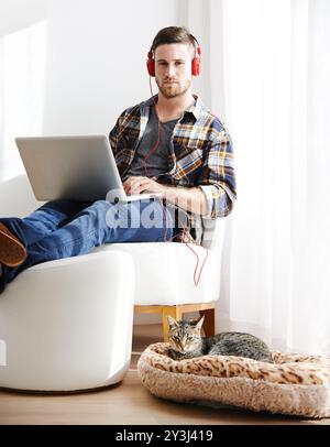 Homme, maison et chat sur ordinateur portable avec casque pour éditer des vidéos en tant que vidéaste avec travail à distance. Personne masculine, employé et sérieux sur portrait comme Banque D'Images