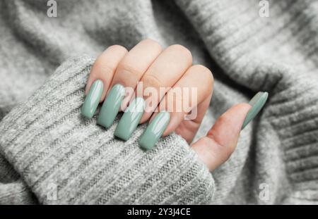 Une main élégante avec des ongles vert menthe bien manucurés est délicatement placée sur un pull gris doux, mettant en valeur un accent à la mode dans un environnement confortable. Banque D'Images