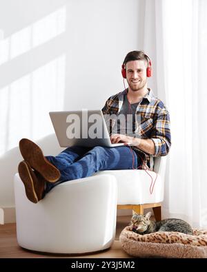 Homme, maison et chat sur ordinateur portable avec casque pour le montage de vidéos en tant que vidéaste avec travail à distance. Personne masculine, animal de compagnie et heureux ou sourire sur portrait comme Banque D'Images