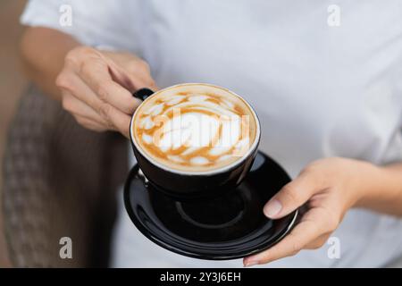 Une personne tenant une tasse noire de café Latte art sur une soucoupe noire, avec un fond flou de sièges extérieurs. Banque D'Images