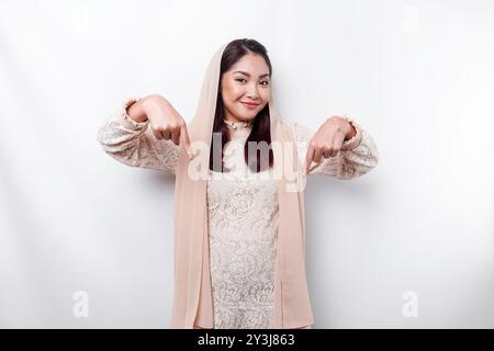 Femme musulmane asiatique excitée hijab pointant vers l'espace de copie en dessous d'elle, isolé par fond blanc. Banque D'Images