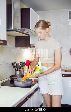 La jeune femme prépare un repas dans une cuisine bien éclairée, ajoutant des légumes frais tranchés dans une poêle sur la cuisinière Banque D'Images