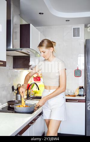 Une jeune femme en tenue décontractée cuisine un plat coloré dans une cuisine élégante et bien éclairée, concentrée sur sa tâche Banque D'Images