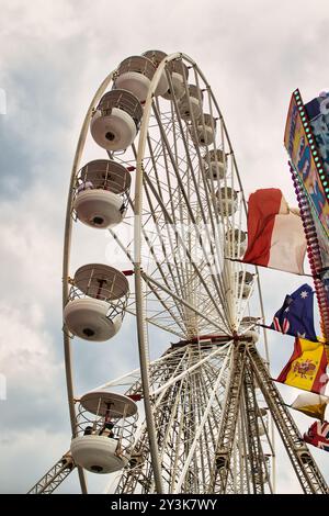 Une vue rapprochée d'une grande roue contre un ciel nuageux, mettant en valeur sa structure et ses gondoles. Des drapeaux colorés de divers pays sont visibles à proximité Banque D'Images