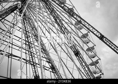 Une vue rapprochée d'une structure de grande roue, mettant en valeur sa structure métallique complexe et ses gondoles dans un ciel nuageux, en noir et blanc. Banque D'Images