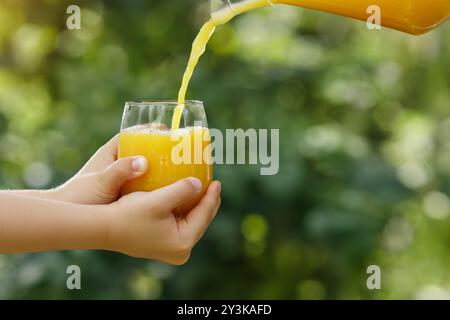 mains d'enfant tenant le verre et le jus d'orange coulant de la cruche avec jardin vert sur le fond Banque D'Images