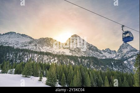 Paysage d'hiver avec le soleil se cacher derrière les Alpes montagnes couvertes par des forêts de sapins, et un cable car crossing. Image capturée à Ehrwald, Austri Banque D'Images