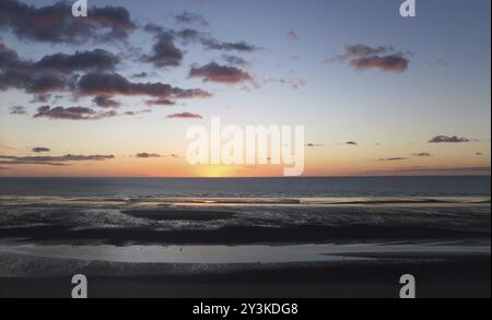 Large scène panoramique de plage au crépuscule avec un ciel de coucher de soleil rose assombrissant et des nuages reflétés dans une mer calme et l'eau sur la plage Banque D'Images