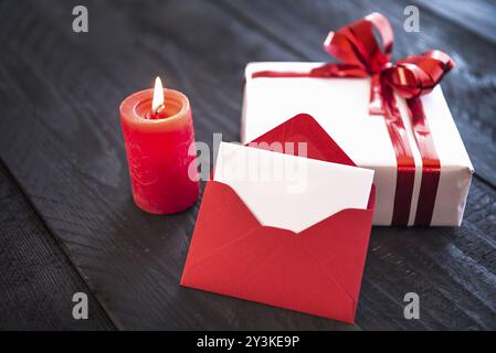 Enveloppe vide rouge appuyée contre un cadeau élégamment emballé, attaché avec un ruban rouge et un noeud, près d'une bougie allumée sur une vieille table en bois Banque D'Images