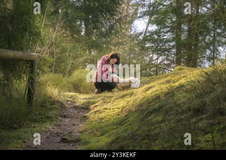 Belle femme brune caressant son chien, sur un chemin, dans une forêt de sapins, sous la lumière du soleil, dans les montagnes de la Forêt Noire, près de la ville de Seebach, Allemagne, UE Banque D'Images