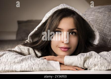 Belle, jeune femme brune, se relaxant sur le canapé, à la maison, vêtue d'un sweat-shirt à capuche confortable, souriant à la caméra Banque D'Images