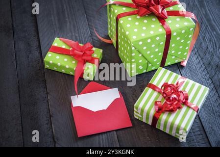 Coffrets cadeaux enveloppés dans du papier vert, attachés avec un ruban rouge et des noeuds et une lettre rouge ouverte, sur une table en bois noir Banque D'Images