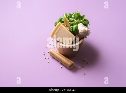 Ingrédients de sauce pesto dans un mortier en bois sur fond violet. Basilic frais, parmesan, ail et pignons de pin pour faire du pesto. Sauce pour pâtes Banque D'Images