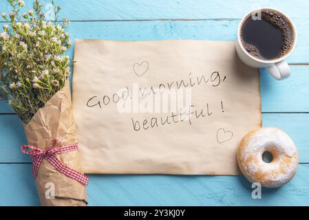 Papier vintage avec le beau message du bon matin et des coeurs, un bouquet de fleurs de printemps, café chaud et beignet, sur un fond bleu en bois Banque D'Images