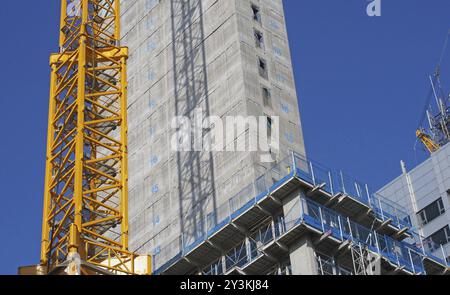 Gros plan d'un grand chantier de construction urbain avec une grue à tour jaune projetant une ombre sur un grand bâtiment en béton et des clôtures de sécurité contre un s bleu Banque D'Images