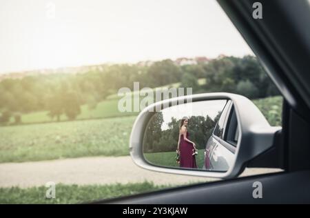 Jeune femme, dans une robe rouge de soirée, avec ses chaussures et un bouquet de fleurs dans ses mains, vue dans le rétroviseur, s'éloignant de la voiture Banque D'Images