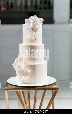 Elégant gâteau de mariage à trois niveaux orné d'orchidées blanches délicates et monogramme d'or sur le stand géométrique moderne. Banque D'Images