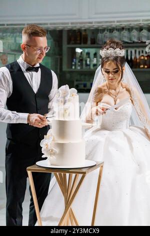 Cérémonie de découpe de gâteau de mariage élégante avec mariée et marié dans une tenue élégante célébrant leur Journée spéciale. Banque D'Images