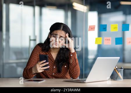 Triste femme d'affaires déçue tenant le téléphone dans les mains, employé de bureau a reçu un message avec de mauvaises nouvelles en ligne, femme latino-américaine mécontente du résultat des affaires à l'intérieur du bâtiment. Banque D'Images