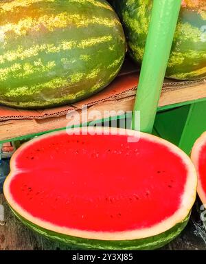 Une demi-pastèque juteuse mûre sur le fond de pastèques entières au marché du fermier. Récoltez des fruits biologiques riches en humidité. Gros plan Banque D'Images